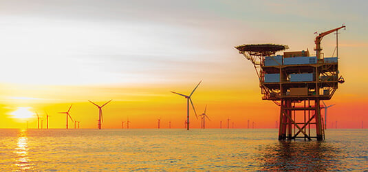 Oil and gas drilling station at sea at sunset with huge wind-turbine farm in background depicts sustainable energy solutions.