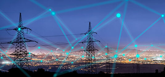 Blue, illuminated electricity grid in front of electrical towers and sprawling city at dusk depicts grid management.