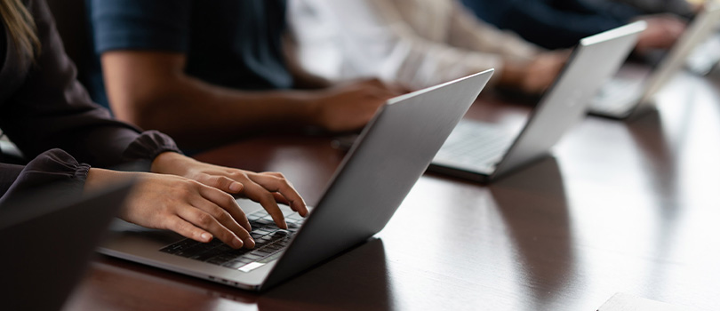 A close-up of a person typing on a laptop at a desk surrounded by other people typing on their laptops.