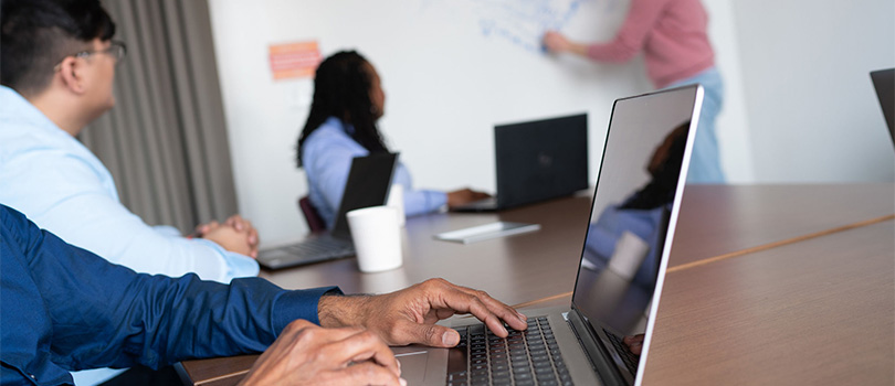 A close-up of a person typing on a laptop keyboard at a desk, surrounded by colleagues all watching an instructor write on a whiteboard.