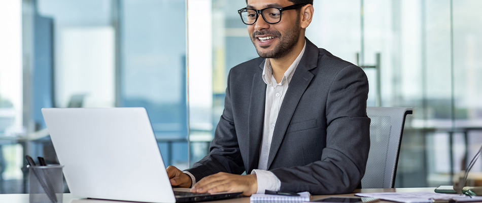 A person with short hair and glasses sitting at a desk working on a laptop in an office environment.