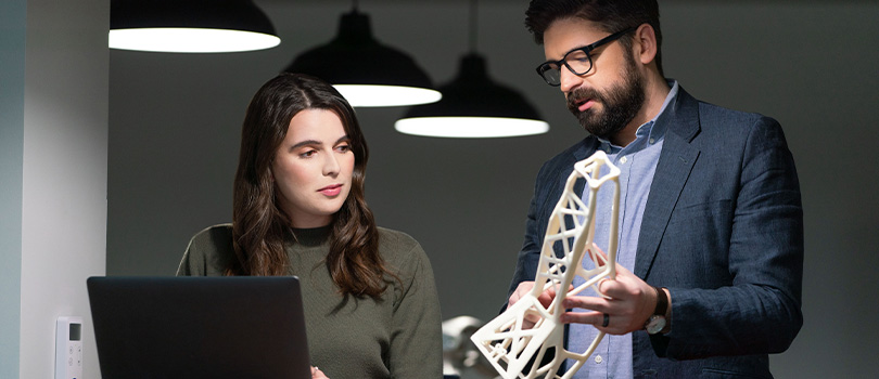 Man showing 3D printed part to woman at laptop computer