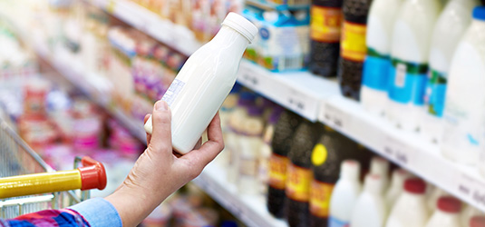 A photo of supermarket shelves in the background and a person's hand holding up a white plastic bottle in the foreground.