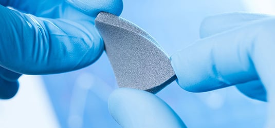 A close-up shot of a small piece of grey ceramic material held by a person wearing blue lab gloves.