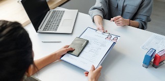 A dark haired woman hands her passport and immigration paperwork to another woman sitting across a table.